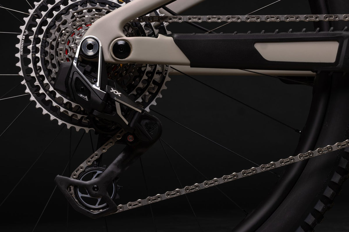 Close-up of a bicycle's rear derailleur and chainring system, showing black and silver components with a beige frame element against a dark background.