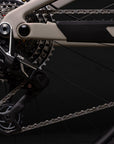 Close-up of a bicycle's rear derailleur and chainring system, showing black and silver components with a beige frame element against a dark background.