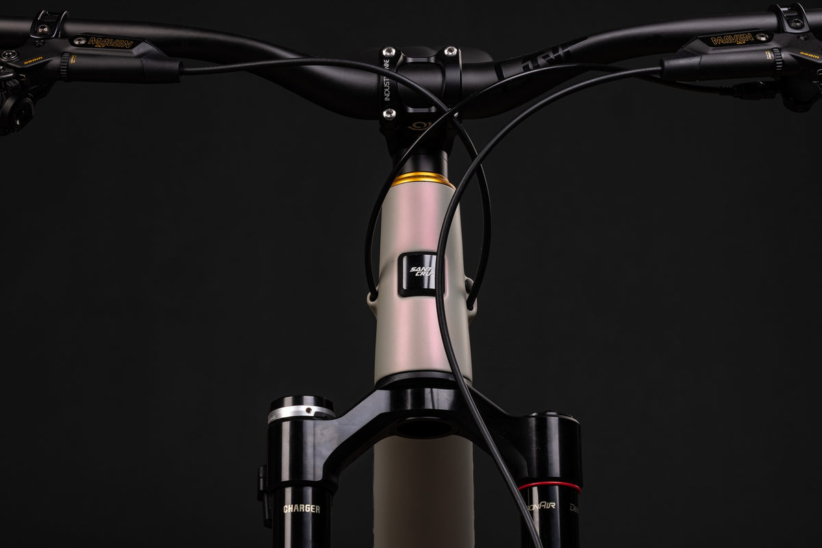 Close-up of a mountain bike handlebar and fork assembly against a dark studio background. The frame is a pale blush pink with a black headtube and black fork, and small gold branding is visible on the handlebars and fork.