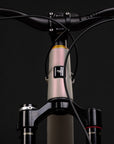 Close-up of a mountain bike handlebar and fork assembly against a dark studio background. The frame is a pale blush pink with a black headtube and black fork, and small gold branding is visible on the handlebars and fork.