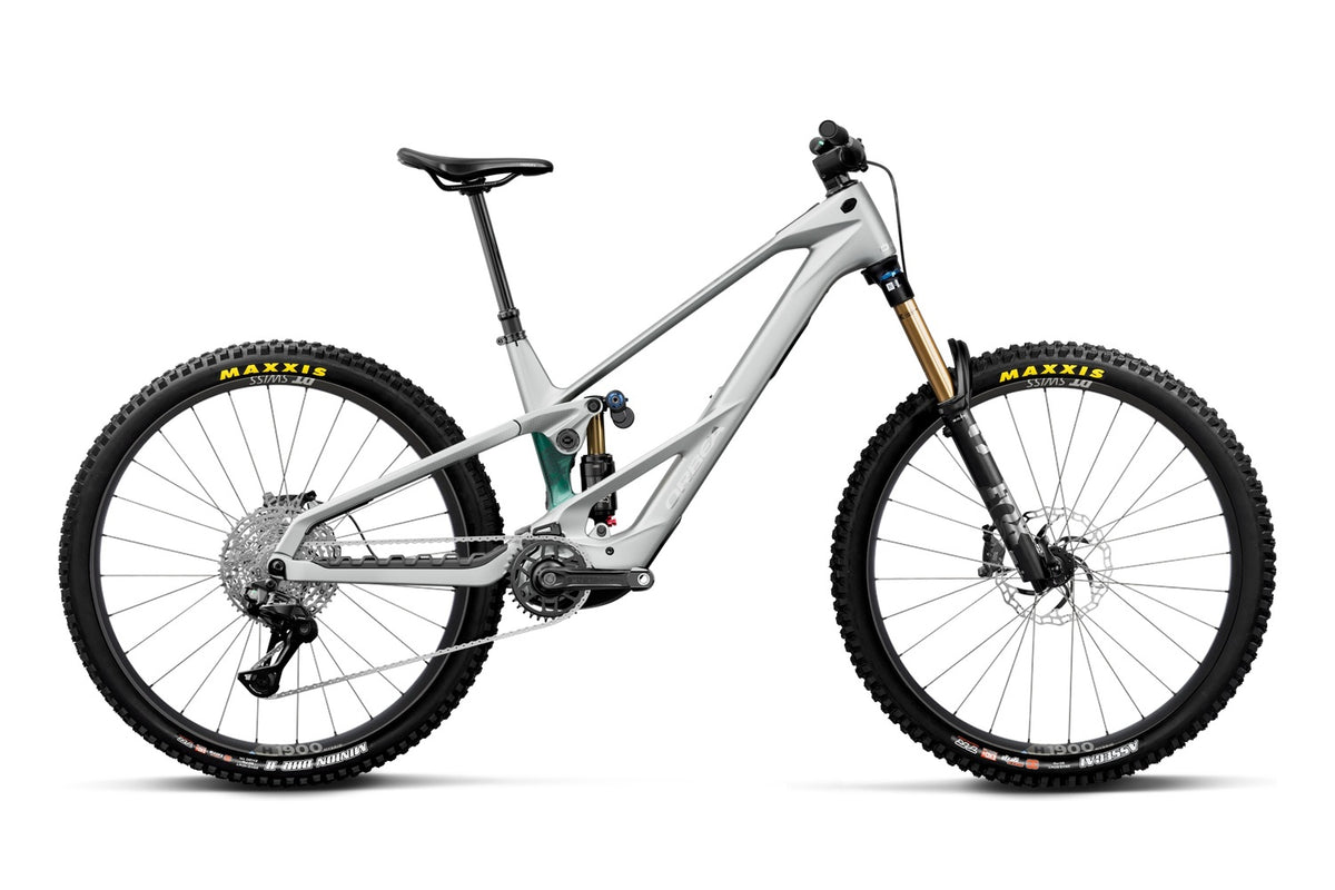 A full-suspension mountain bike in a matte light gray frame with black tires, a black saddle, and gold Fox suspension forks, shown from a side profile on a white background.