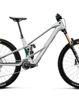 A full-suspension mountain bike in a matte light gray frame with black tires, a black saddle, and gold Fox suspension forks, shown from a side profile on a white background.