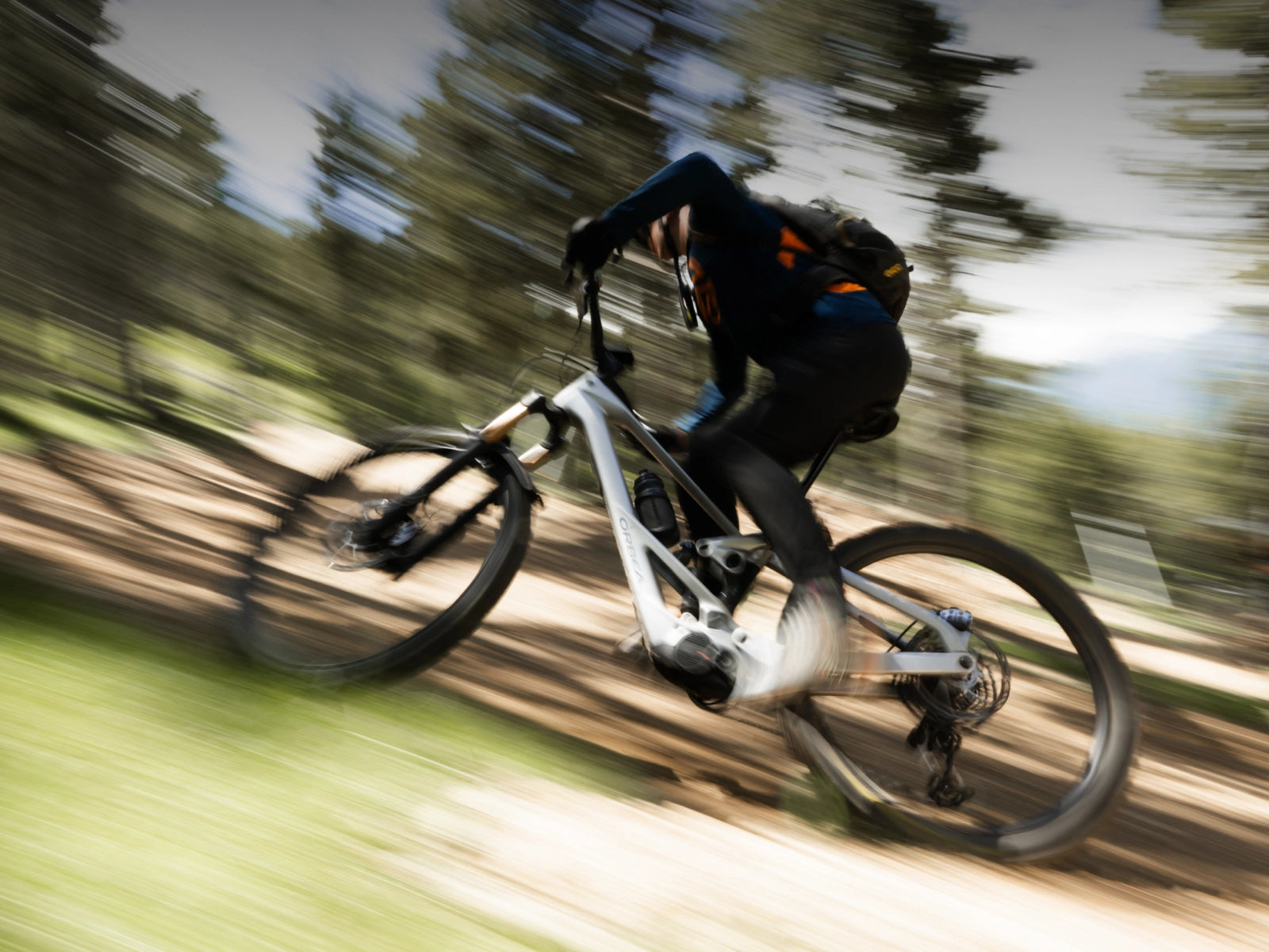 A mountain biker in black clothing rides a white full-suspension electric mountain bike on a dirt trail, captured with a dynamic motion blur effect.